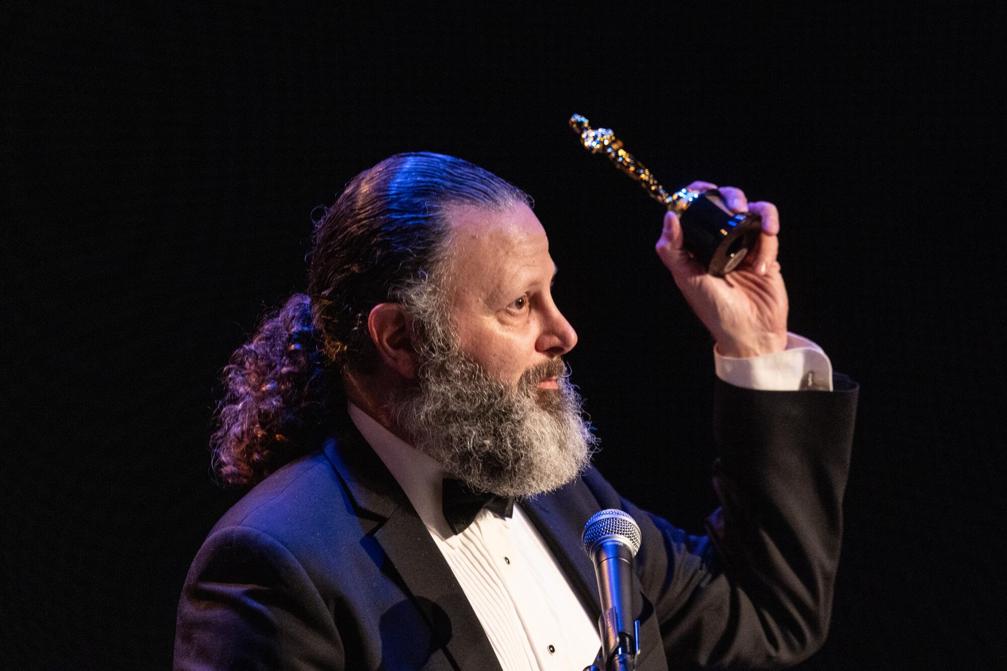 A man with a graying beard, in a tuxedo, holds up a statuette while speaking at a mic