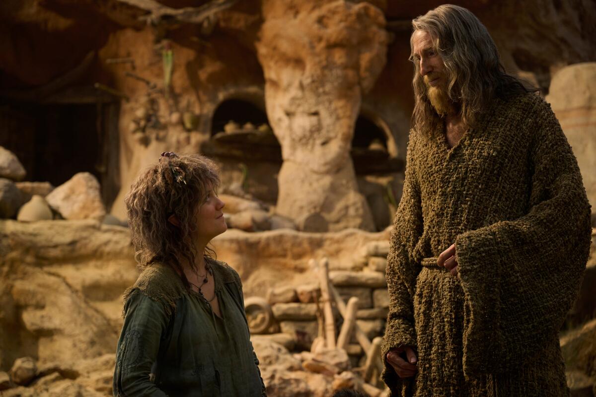 A woman with shaggy hair and green robe looks up at a tall man with long gray hair and a beard.