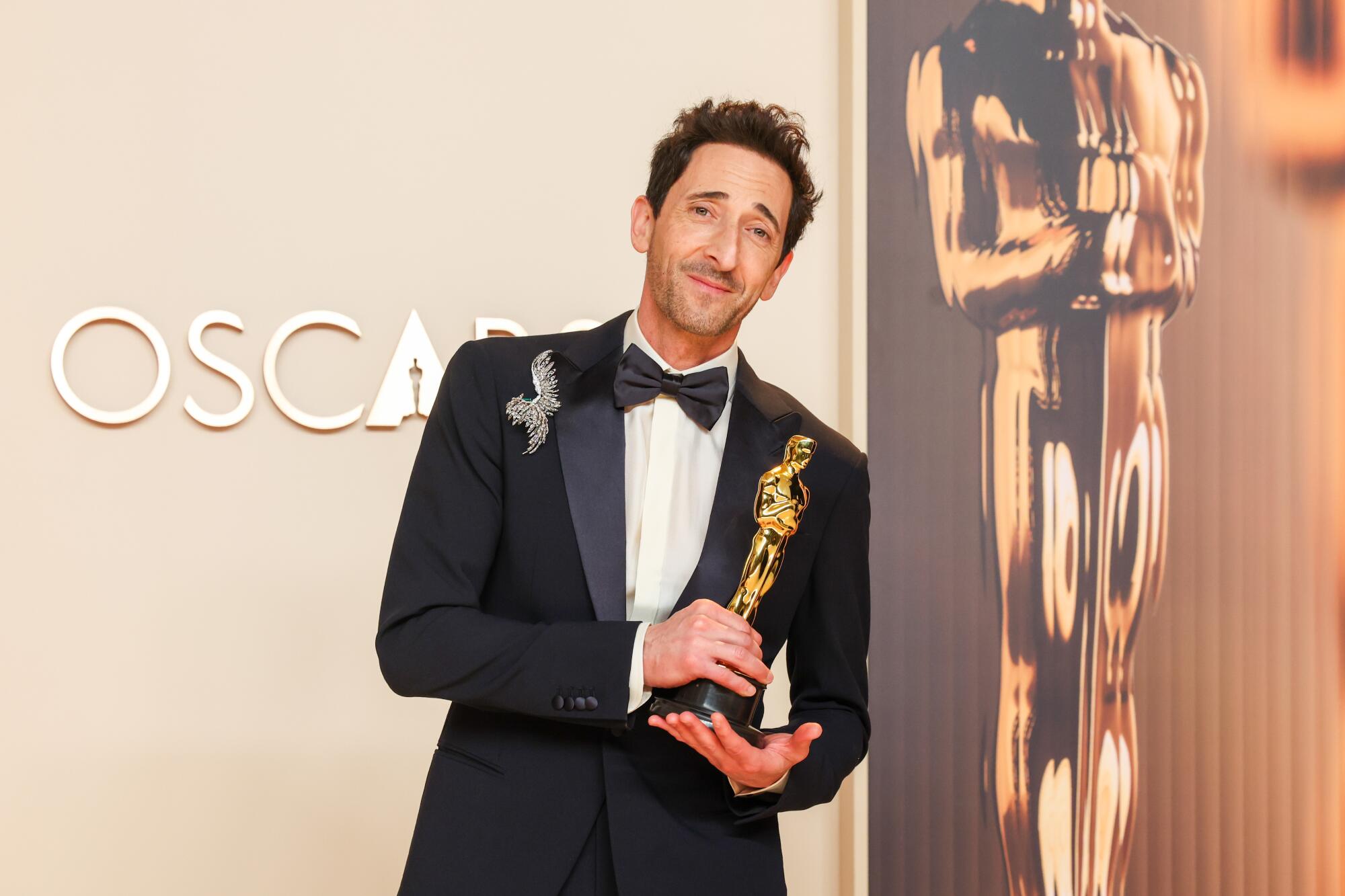 Adrien Brody in a tuxedo leans to his left and holds his Oscar statuette in his hands. He stands in front of a gold backdrop