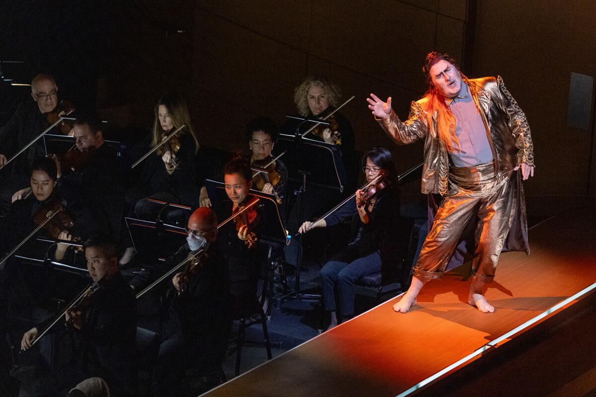 A costumed singer in a golden suit performs on a catwalk above string players of the L.A. Phil during "Das Rheingold."