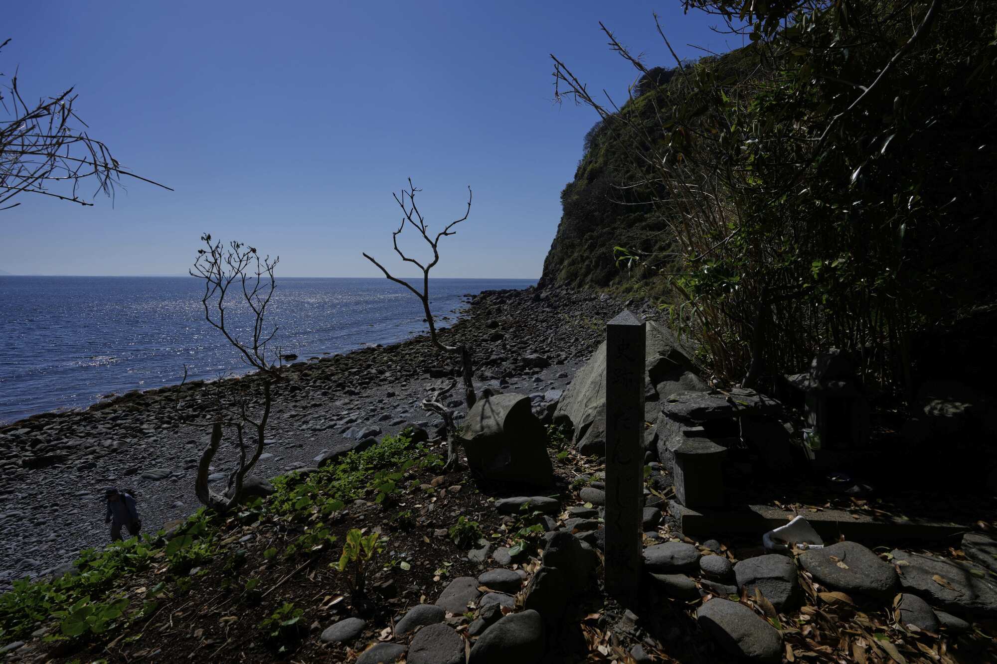 A fisherman walks on a seashore near a grave marker.