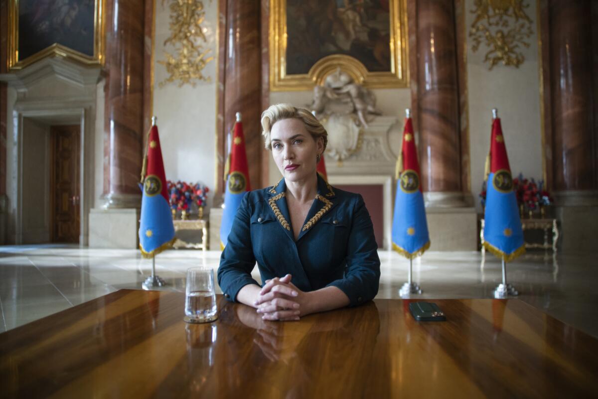 A woman in a blue suit sits at a wooden table in an ornate palace.
