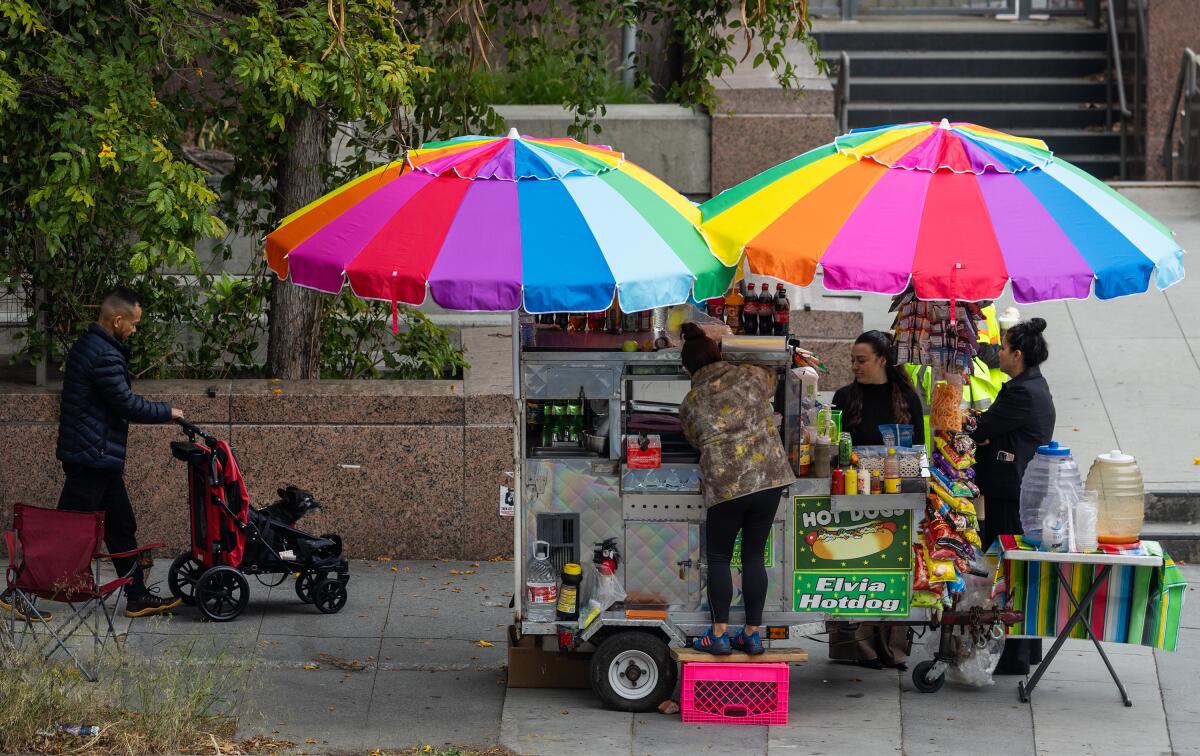 Brightly colored umbrellas shelter a food cart and its operator from the sun.