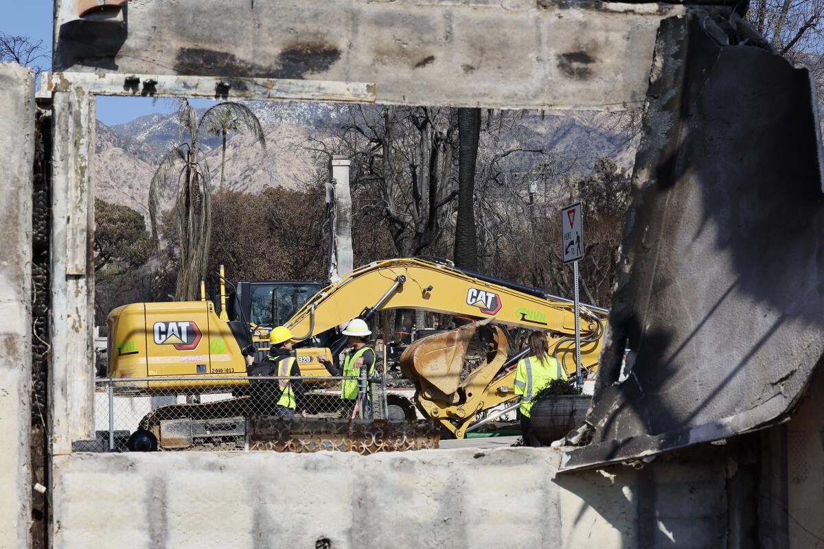 A yellow CAT excavator is seen through the window of a burned wall, with mountains and scorched palm trees in the distance