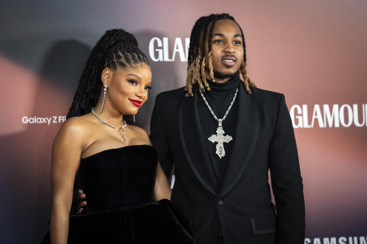 Halle Bailey standing in a black strapless dress next to DDG in a dark suit and large cross necklace