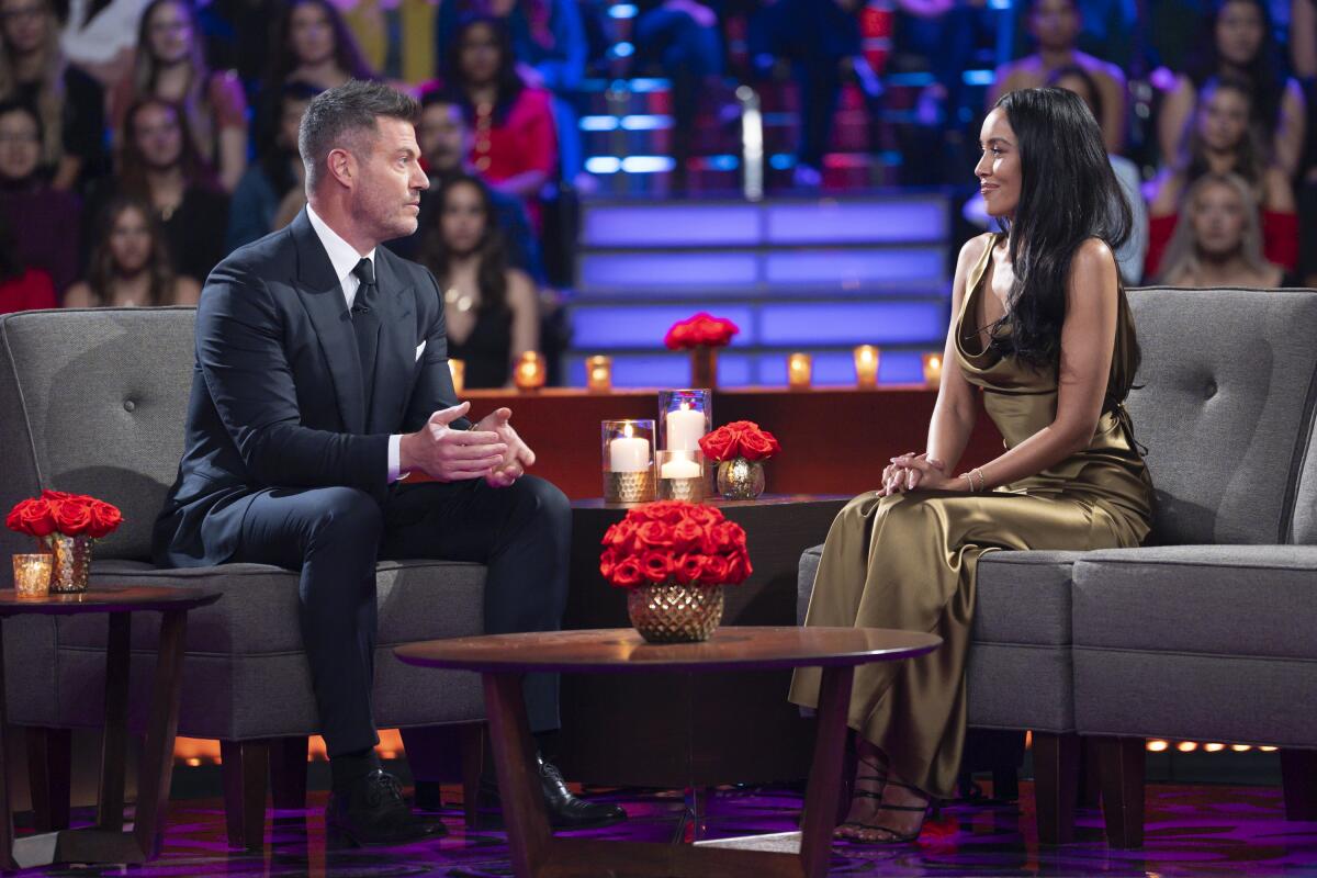 A man in a dark suit and tie sitting across from a woman with long dark hair in a golden sleeveless gown.