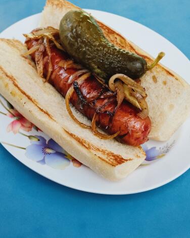 A large sausage in a bun topped with grilled onions and a pickle on a blue table at Stara Pierogi and Sausage