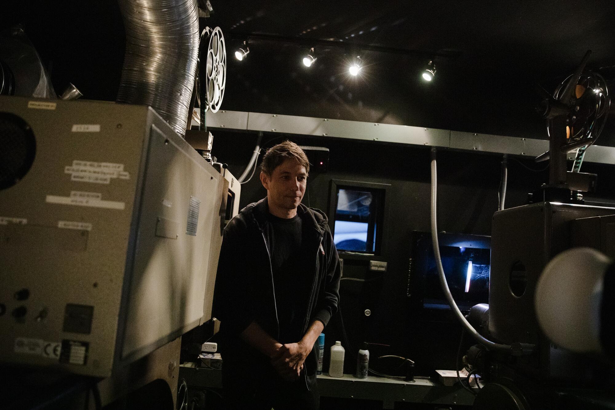 A man in a dark hoodie stands next to a film projector.