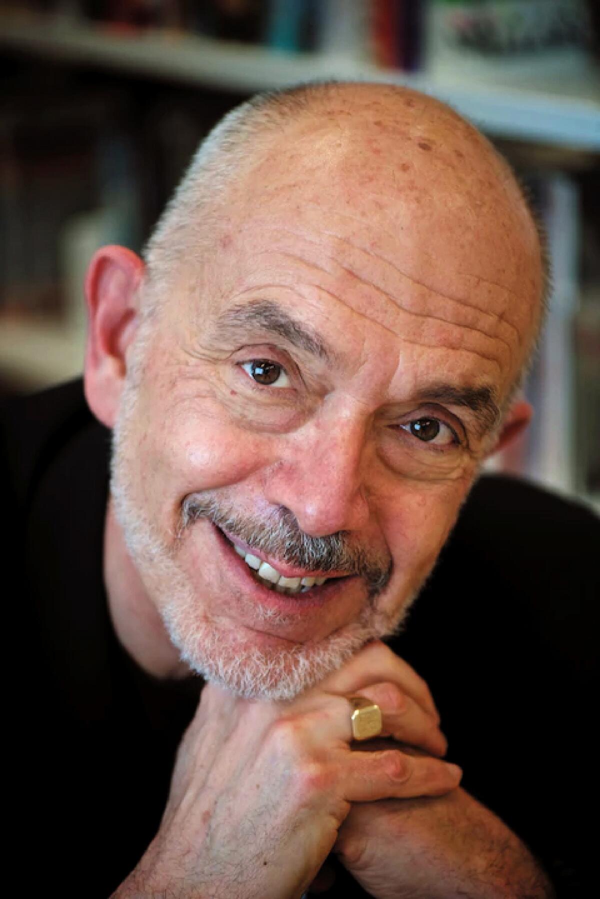 A man with white hair, wearing a gold ring, smiles at the camera.