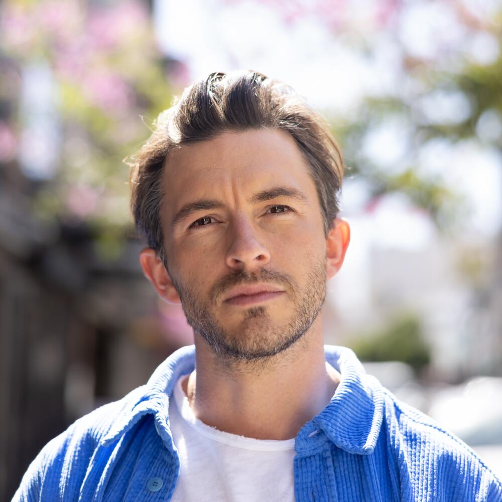 A man in a blue shirt over a white T-shirt, standing before a flowering tree.