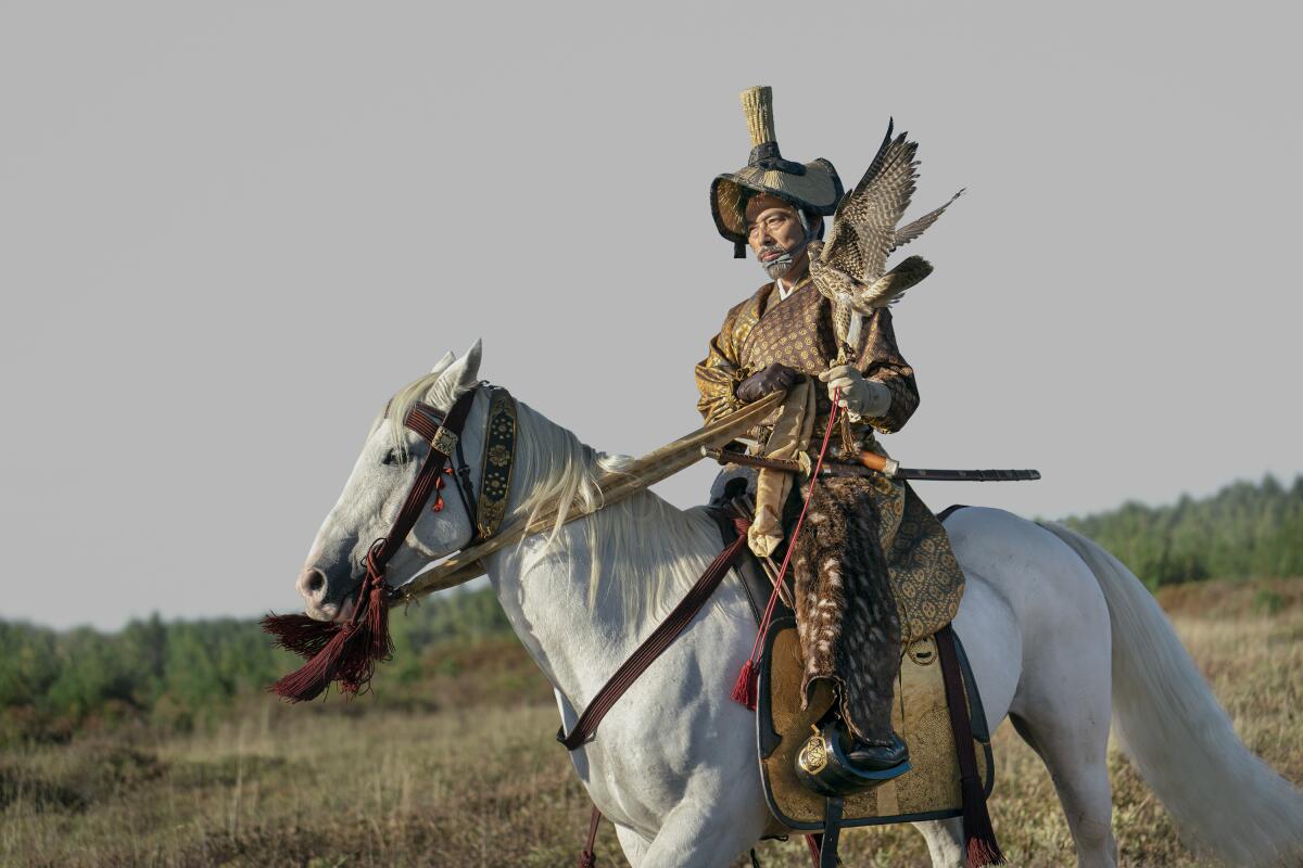 A man in samurai gear sits atop a white horse with a falcon perched on his hand.