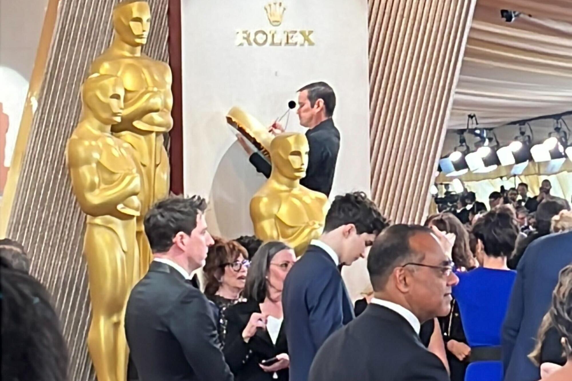 A man, standing beside life-size Oscar statues, fixes a large clock under a Rolex logo.