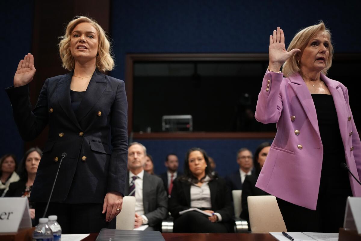 A woman in a dark suit and a woman in a pink blazer raise their right hands as they stand in front of a long table.