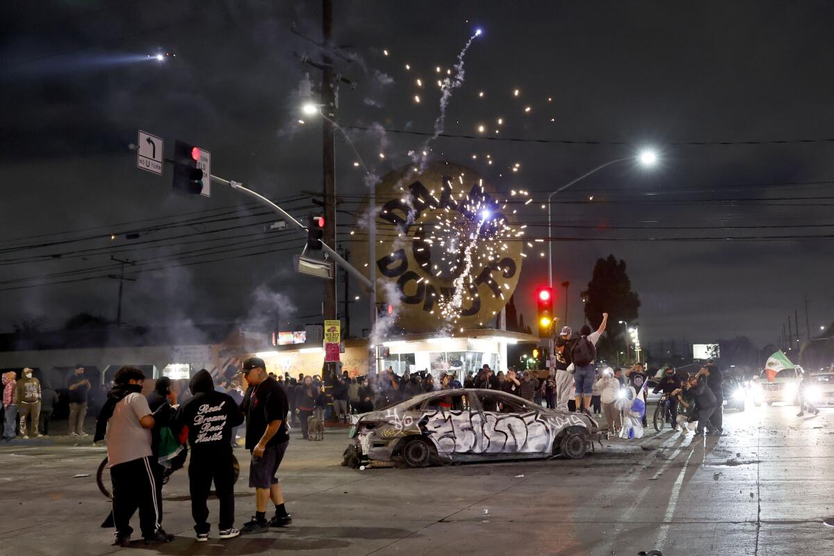 Protesters explode fireworks over a torched car at an intersection in Compton at night.