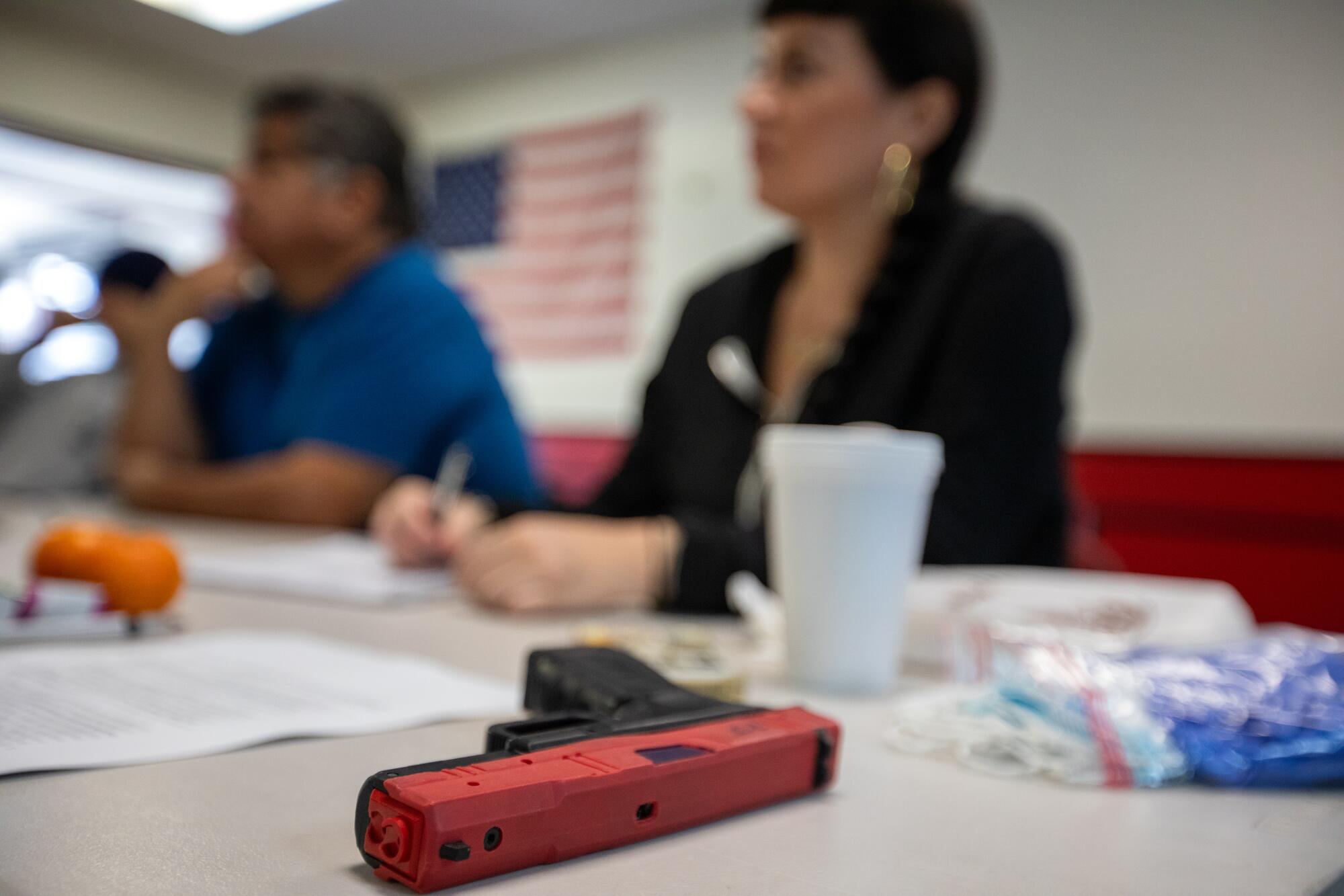 A prop Glock pistol with a red slide resting on a table where two students are seated