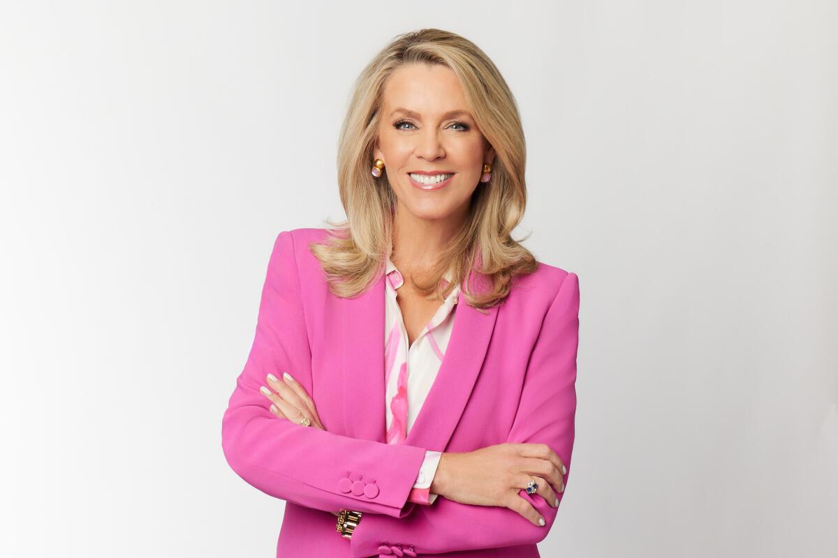 Deborah Norville stands in a pink suit with her arms crossed in front of her