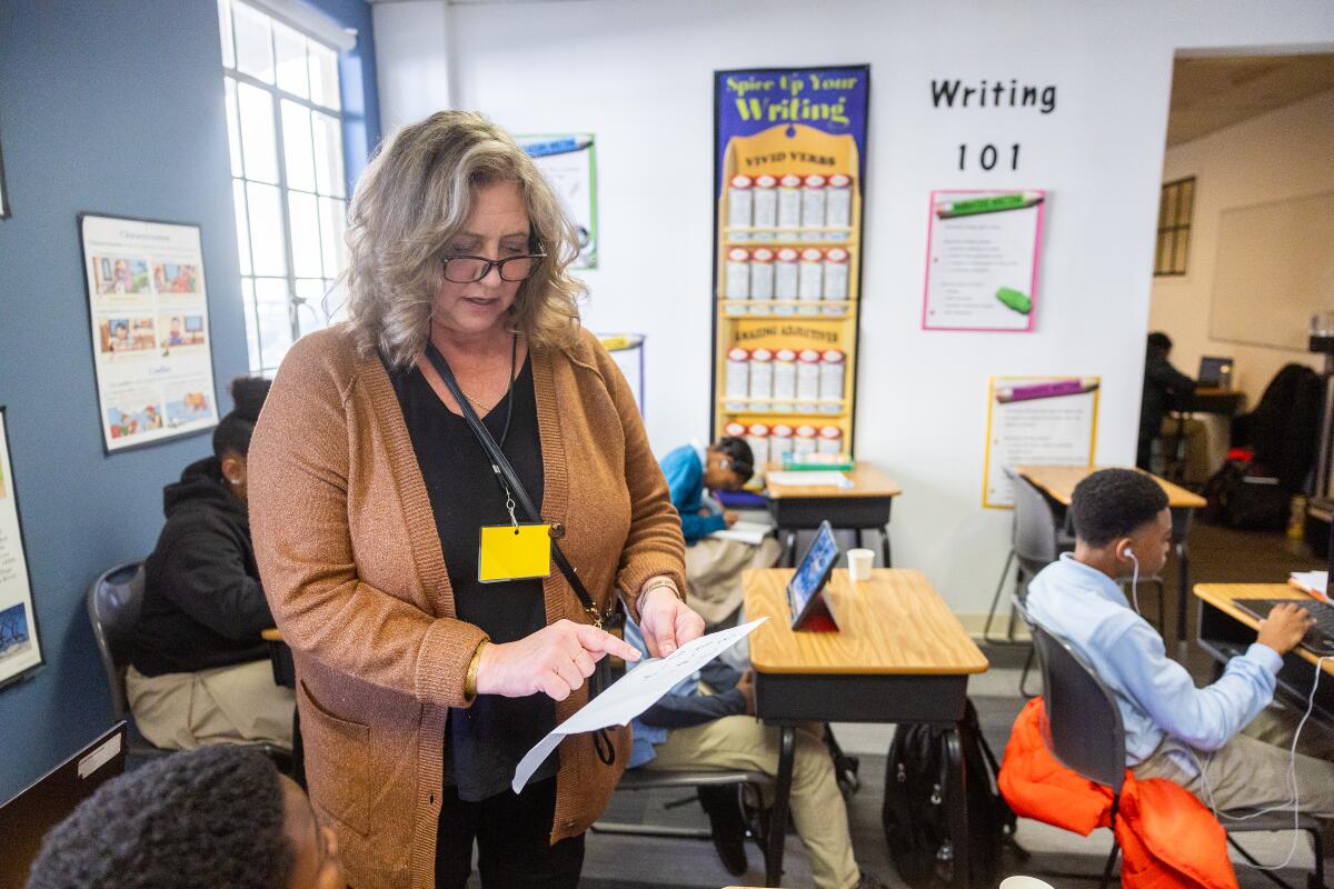 A teacher points at a piece of paper while standing in a classroom full of children.